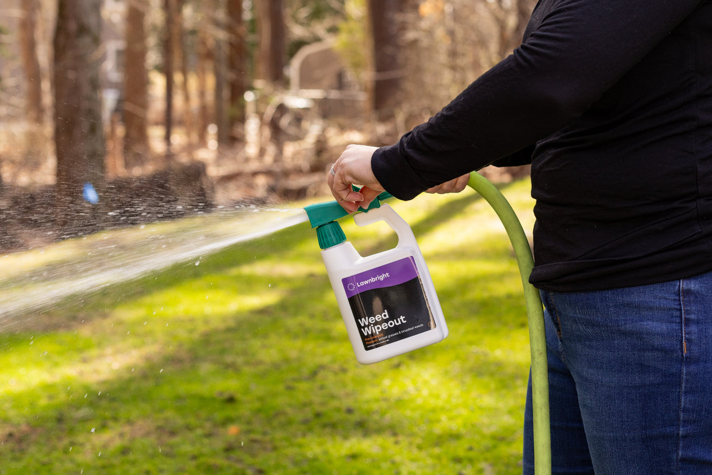 Person holding a bottle labeled 'Weed Out' over a garden hose with a grassy area and trees in the background.